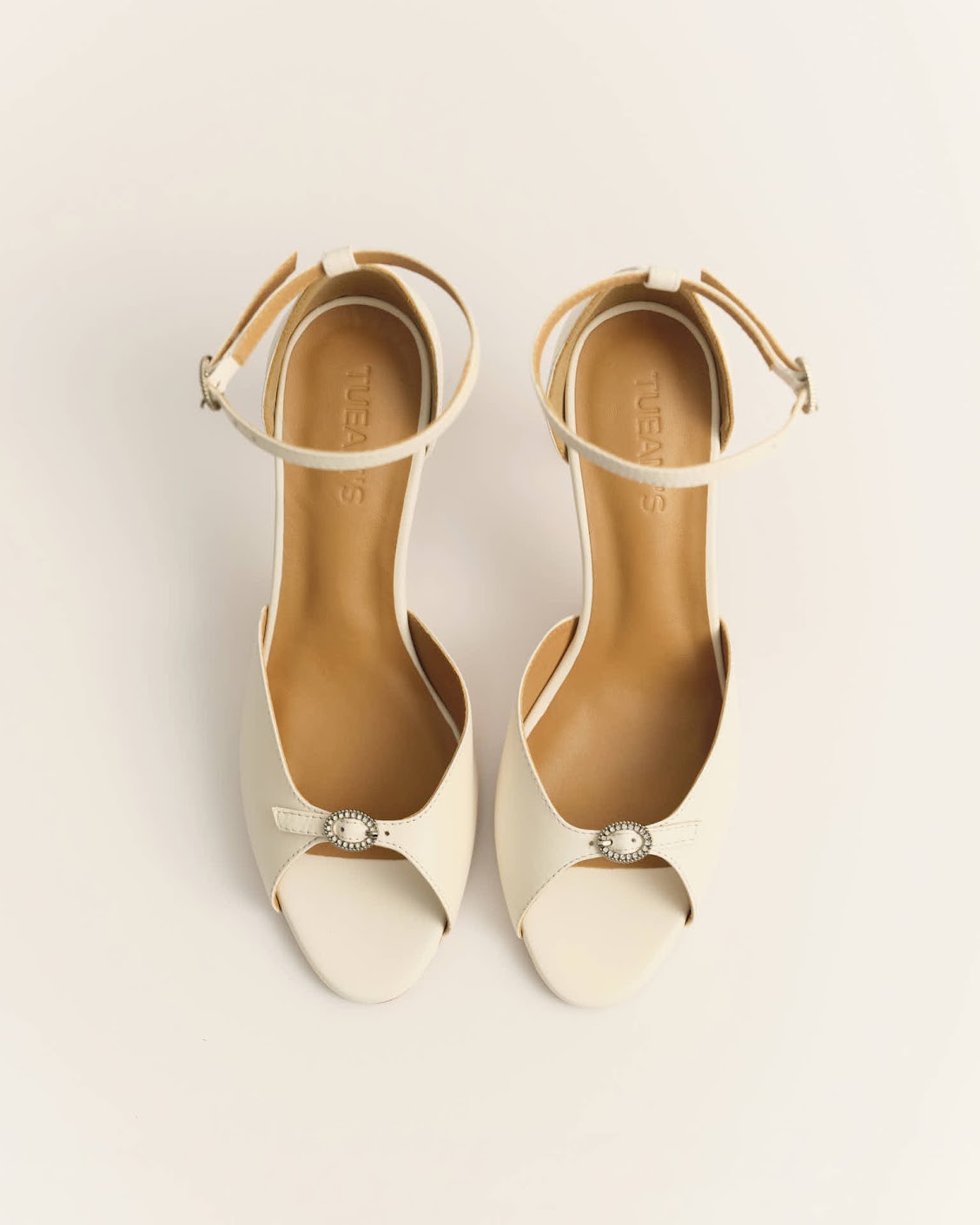 Cream open-toe high-heeled sandals with ankle straps and a jeweled circular ornament at the front.