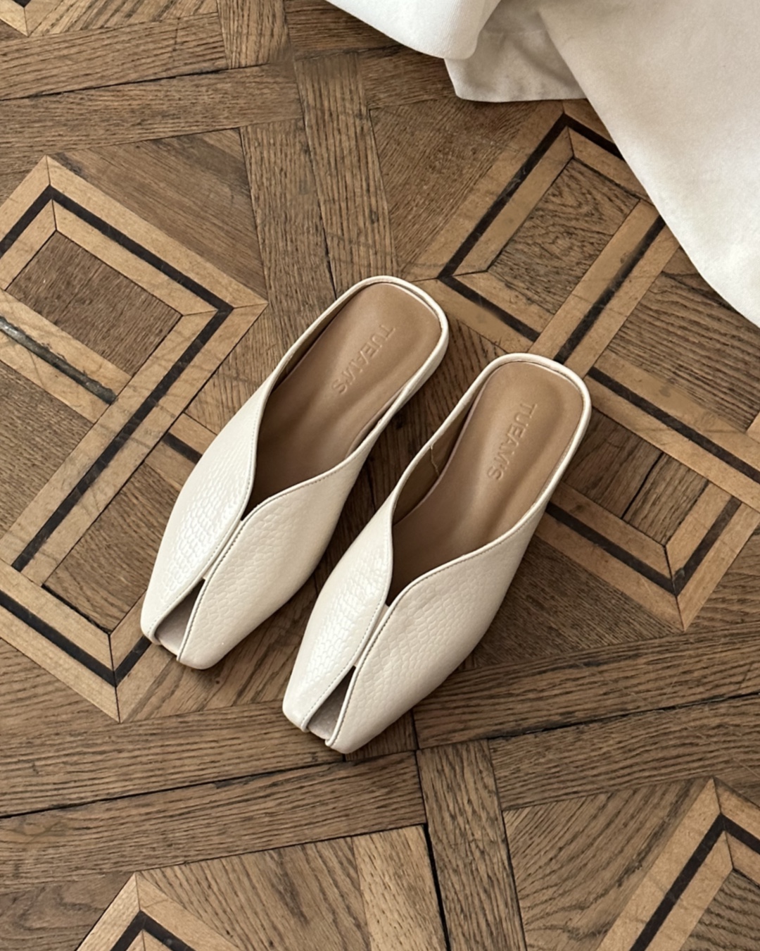 Pair of white textured slip-on mules with pointed toes on a wooden parquet floor, seen from above.