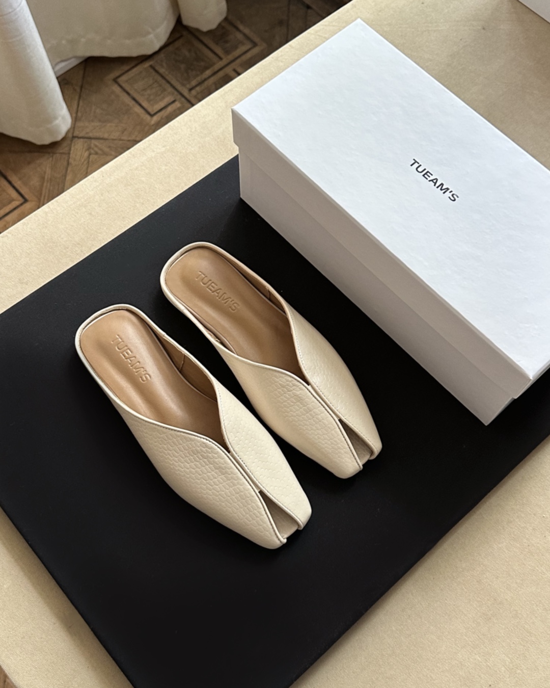 Cream peep-toe slip-on mules in textured beige next to a white TUEAM'S shoebox on a black display surface.