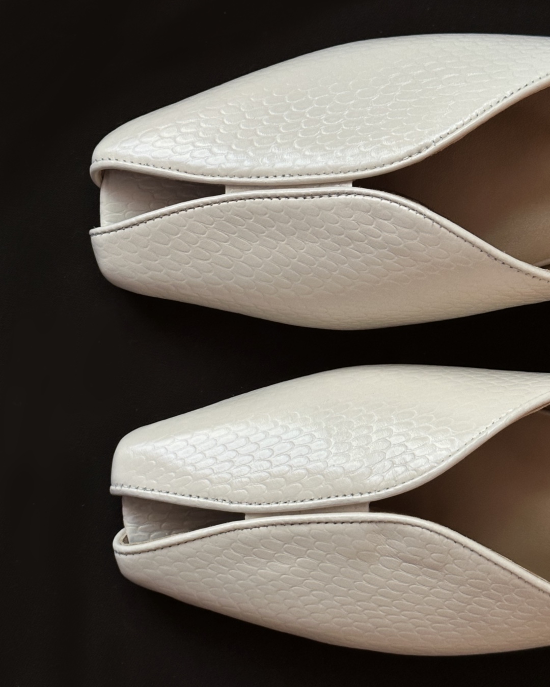White textured leather pointed-toe shoes with curved seams set against a dark background (close-up of the top portions).