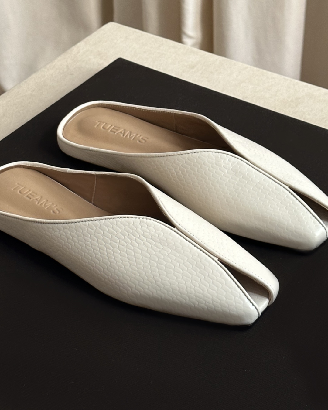 White textured slip-on mules with tan insole resting on a dark surface, showing a chic open-back design.
